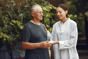 an elderly man is stood outside by a large bush. a nurse is stood next to him. she has her hand on his shoulder, and is passing him a sleeve of tablets in the other. she is looking down at his hand. he is looking off into the distance. they are both smiling.