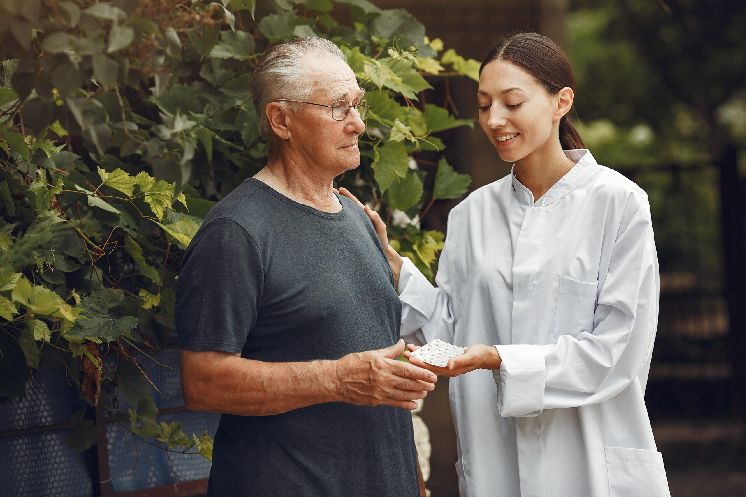 an elderly man is stood outside by a large bush. a nurse is stood next to him. she has her hand on his shoulder, and is passing him a sleeve of tablets in the other. she is looking down at his hand. he is looking off into the distance. they are both smiling.