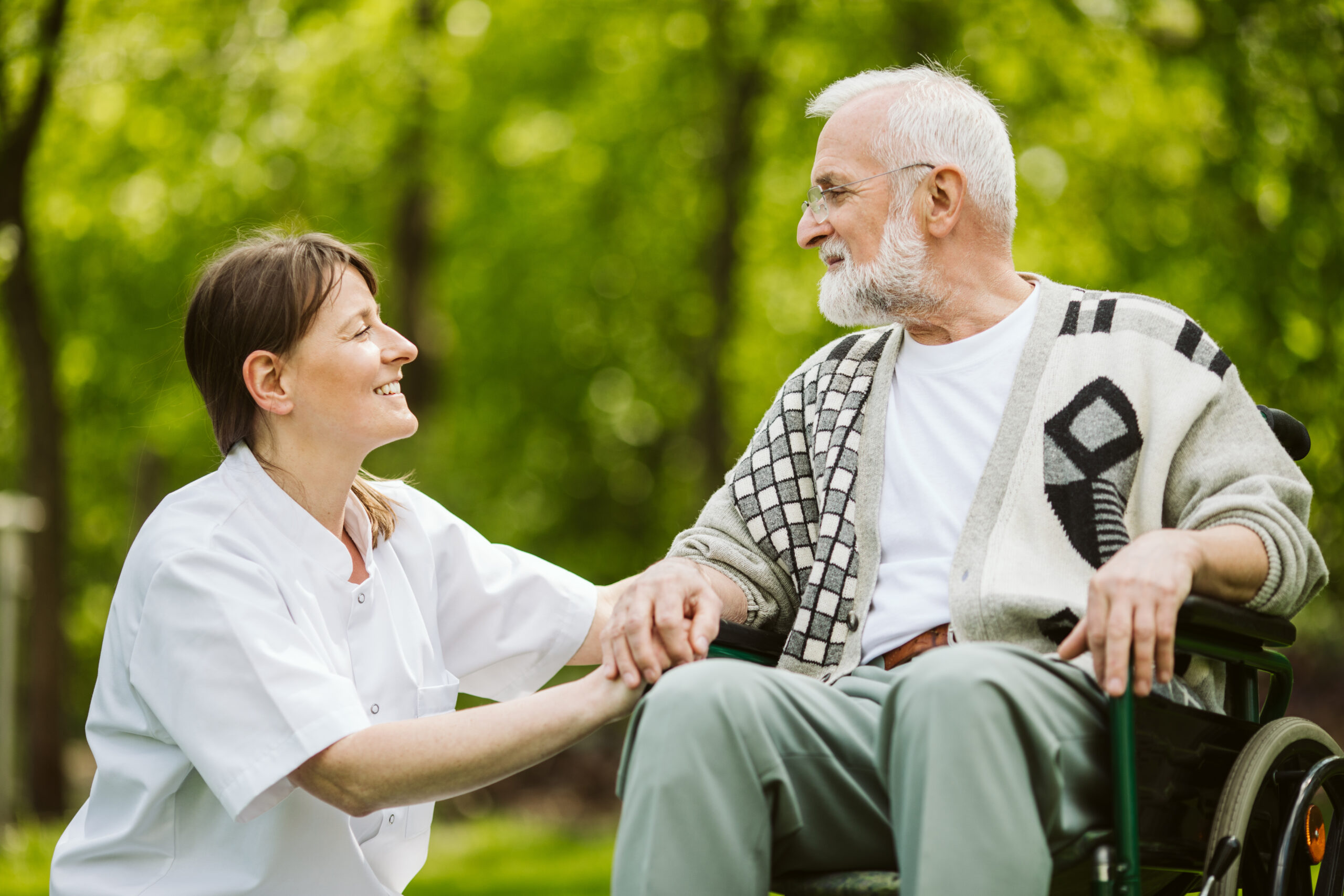 an elderly man is sat outside in a wheelchair. a nurse is knelt down next to him, holding his hand. they are both looking at each other and smiling.