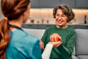 an elderly woman and a nurse are indoors. the elderly woman is sat on a sofa, and is holding a spiky orange ball in her hand. the nurse is holding one of her hands, and supporting the one with the ball. they are both looking at each other. the elderly woman is smiling.
