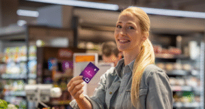 a woman is in a grocery store. she is in focus. she is looking forwards and smiling. in her hand she is holding a purple and pink gradient card with the money carer logo on.