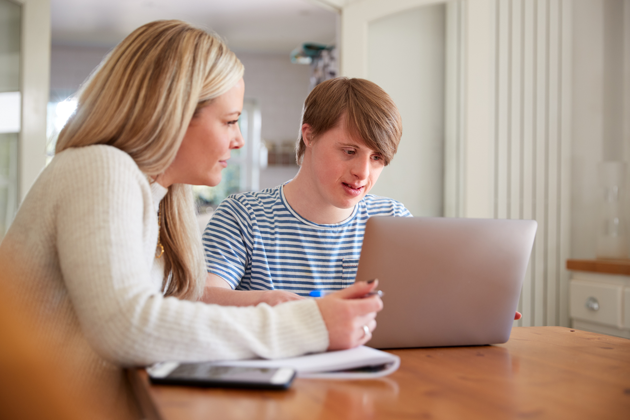 a young man and his mother are sat at a table indoors. the mother, on the left, has an open notebook and a phone in front of her, and a pen in her hand. the young man, on the right, has a laptop open in front of him. they are both looking at the laptop screen. they look concentrated.