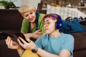 a young person and their mother are in the living room. the mother is lying down on the sofa and looking over their shoulder at a phone screen. the young person has headphones on and is sat on the floor next to the sofa. they are holding the phone towards their mother.