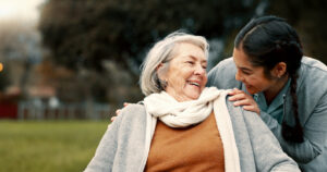 an elderly woman is sat down outside. a younger woman is looking at her and has her hands around the other woman's shoulders. they are both smiling at each other.