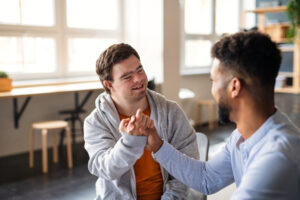 two men are sat indoors. they are holding hands. the face of the man on the right is obscured. the man on the right has down-syndrome. he is looking at the other man and smiling.