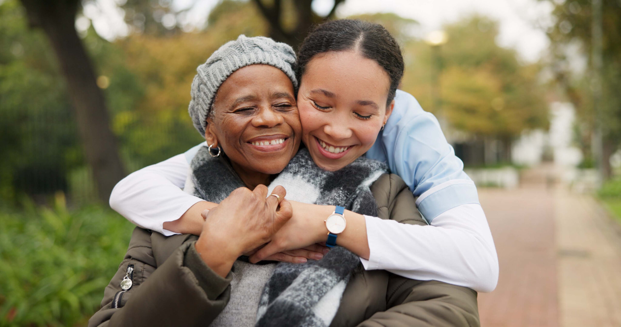 an elderly woman is at down outside with a hat on. her granddaughter is stood behind her and is leaning down to hug her. they are both smiling.