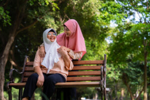 an elderly woman wearing a white headscarf is sat outdoors on a bench, on a sunny day. she is looking up to the sky and smiling. a younger woman with a pink headscarf is stood behind the bench with her arms around the other woman. the elderly woman has a hand over the younger woman's. the younger woman is looking down at her and smiling.