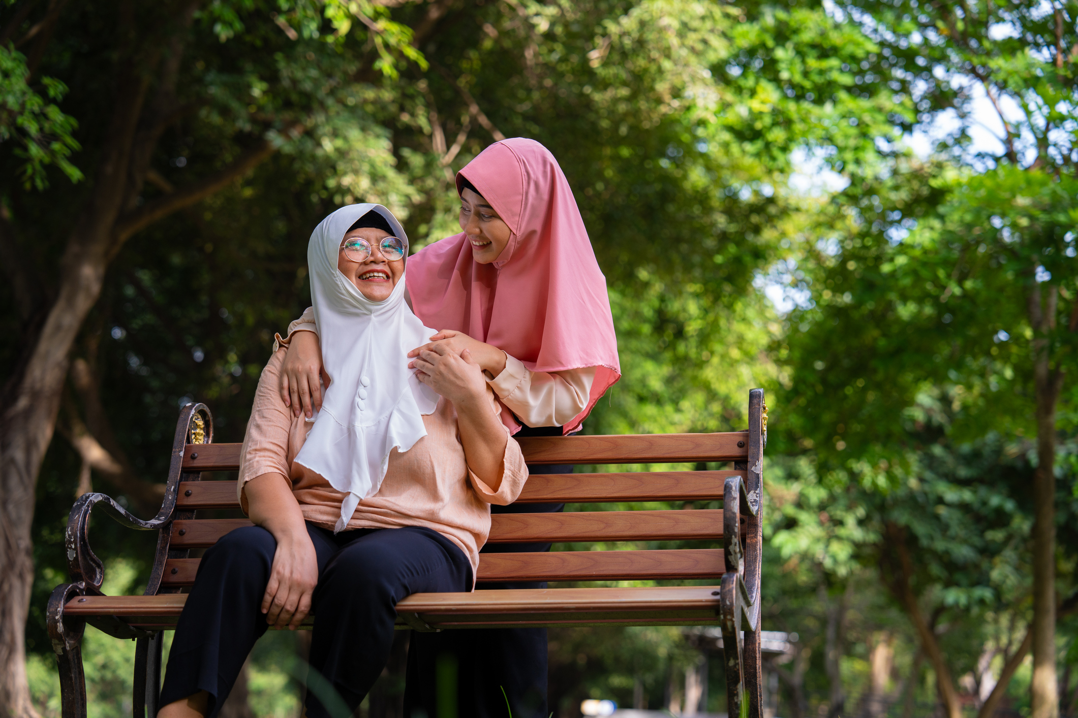 an elderly woman wearing a white headscarf is sat outdoors on a bench, on a sunny day. she is looking up to the sky and smiling. a younger woman with a pink headscarf is stood behind the bench with her arms around the other woman. the elderly woman has a hand over the younger woman's. the younger woman is looking down at her and smiling.