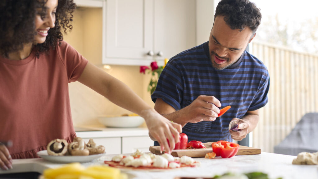 A couple are cooking in a kitchen - the man has down syndrome. They are both smiling. There are ingredients in front of them such as Peppers and Mushrooms.