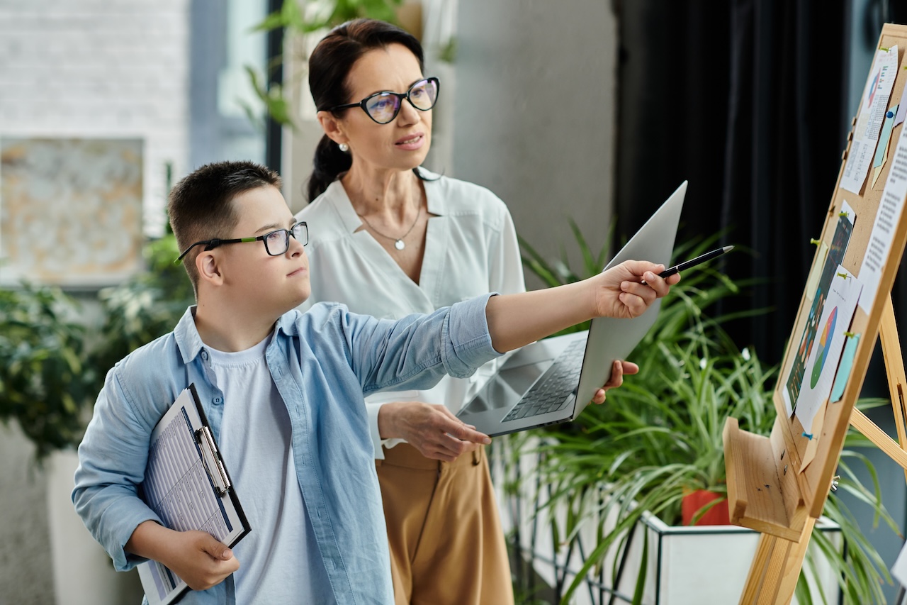 a young man and a woman are indoors in an office environment. they both have glasses on. the young man has down-syndrome. he is holding a clipboard and pointing with a pen at a cork-board full of images. the woman is holding a laptop and looking in the same direction. they both look concentrated.