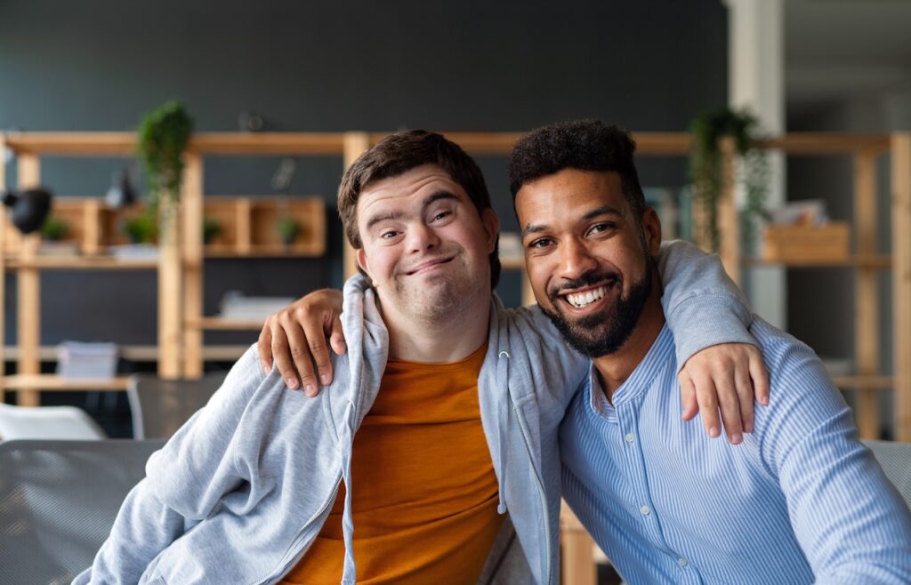 two men are sat indoors on a sofa. they both have an arm around each other. the man on the left has down-syndrome. they are both looking forwards and smiling.
