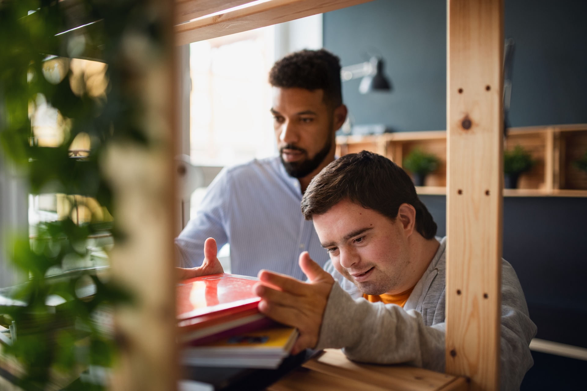 two men are stood indoors by a book shelf. the man on the left looks concentrated. the man on the right has down-syndrome. he is placing a book on top of a pile on the shelf. he looks happy.