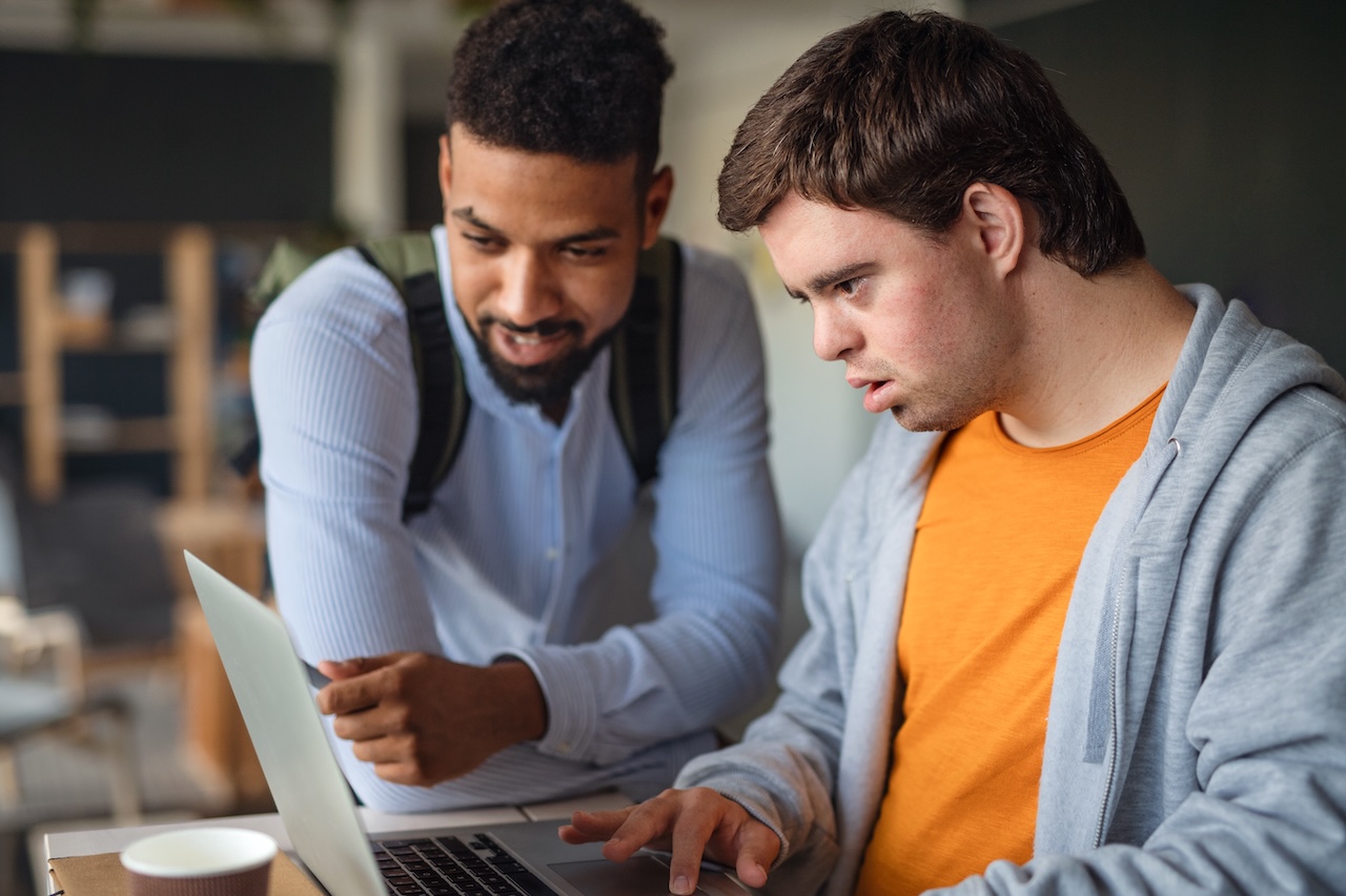 a young man with down-syndrome is looking down at a laptop. there is another man to his left. he is pointing at the screen, and seems to be explaining something. the young man on the right looks concentrated.