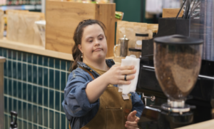 a young woman with down-syndrome is working in a coffee shop. she is stood in front of a coffee machine and holding a folded paper towel. she is wearing an apron and looks concentrated.