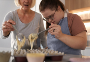 a young woman with down-syndrome and her mother are baking. they are both holding spoons and concentrating on putting cake batter into a baking tin. they both look concentrated.