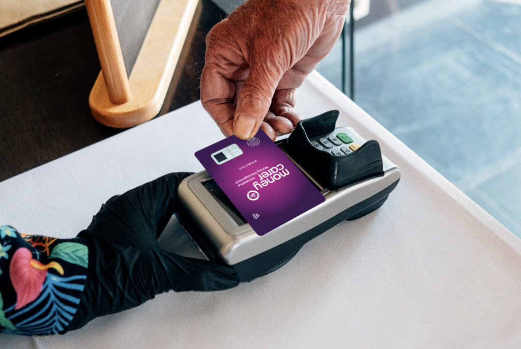 someone is tapping a money carer card to pay by contactless at a card machine. the card is a purple and pink gradient with the money carer logo on it.