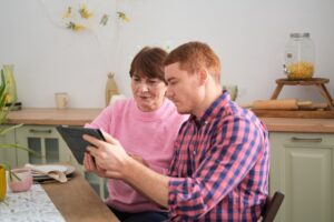 a young man and his mother are sat at a kitchen table. they are both wearing pink. the man, on the right, is holding a digital tablet and angling it towards his mother. the mother is looking at the tablet. they both look concentrated.