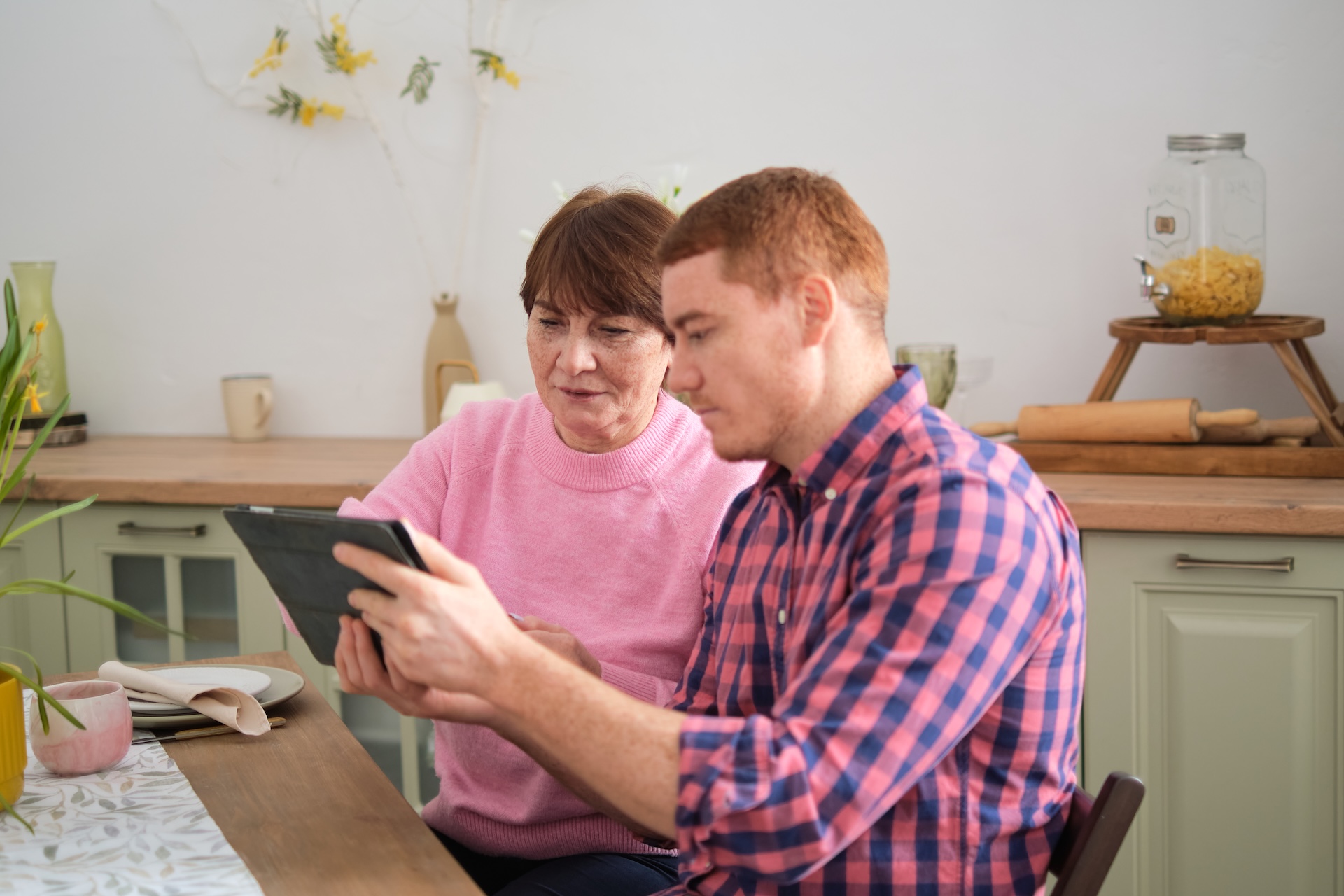 a young man and his mother are sat at a kitchen table. they are both wearing pink. the man, on the right, is holding a digital tablet and angling it towards his mother. the mother is looking at the tablet. they both look concentrated.