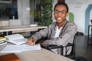 a young man in a wheelchair is sat at a desk indoors. in front of him there is a stack of books and two notebooks. he is writing in the notebook closest to him. he is looking forwards and smiling.