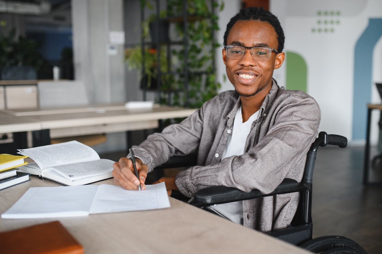 a young man in a wheelchair is sat at a desk indoors. in front of him there is a stack of books and two notebooks. he is writing in the notebook closest to him. he is looking forwards and smiling.