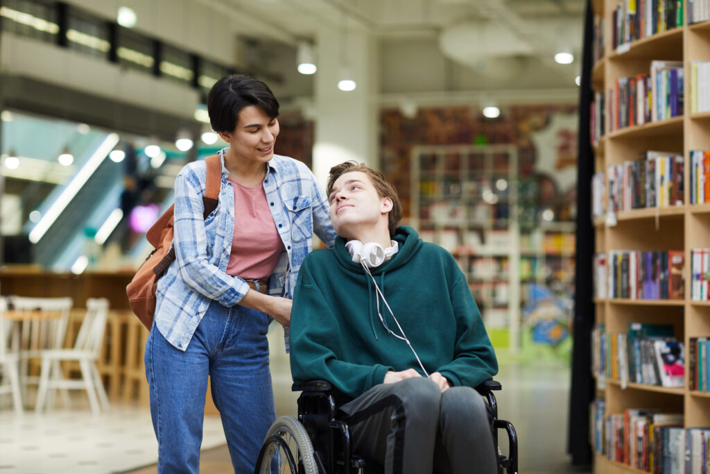 a woman is holding the back of a wheelchair that a young man is using. they are looking at each other and smiling. they appear to be indoors in a library. the young man has headphones around his neck.