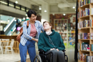 a woman is holding the back of a wheelchair that a young man is using. they are looking at each other and smiling. they appear to be indoors in a library. the young man has headphones around his neck.