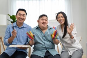 a young man with down syndrome is sat between his parents. the father, on the left, has paint and paintbrushes in his hands. the mother, on the right, is waving. the young man in the middle has his hands painted in a striped pattern of blue, red, green and yellow. they are all looking forward and smiling.