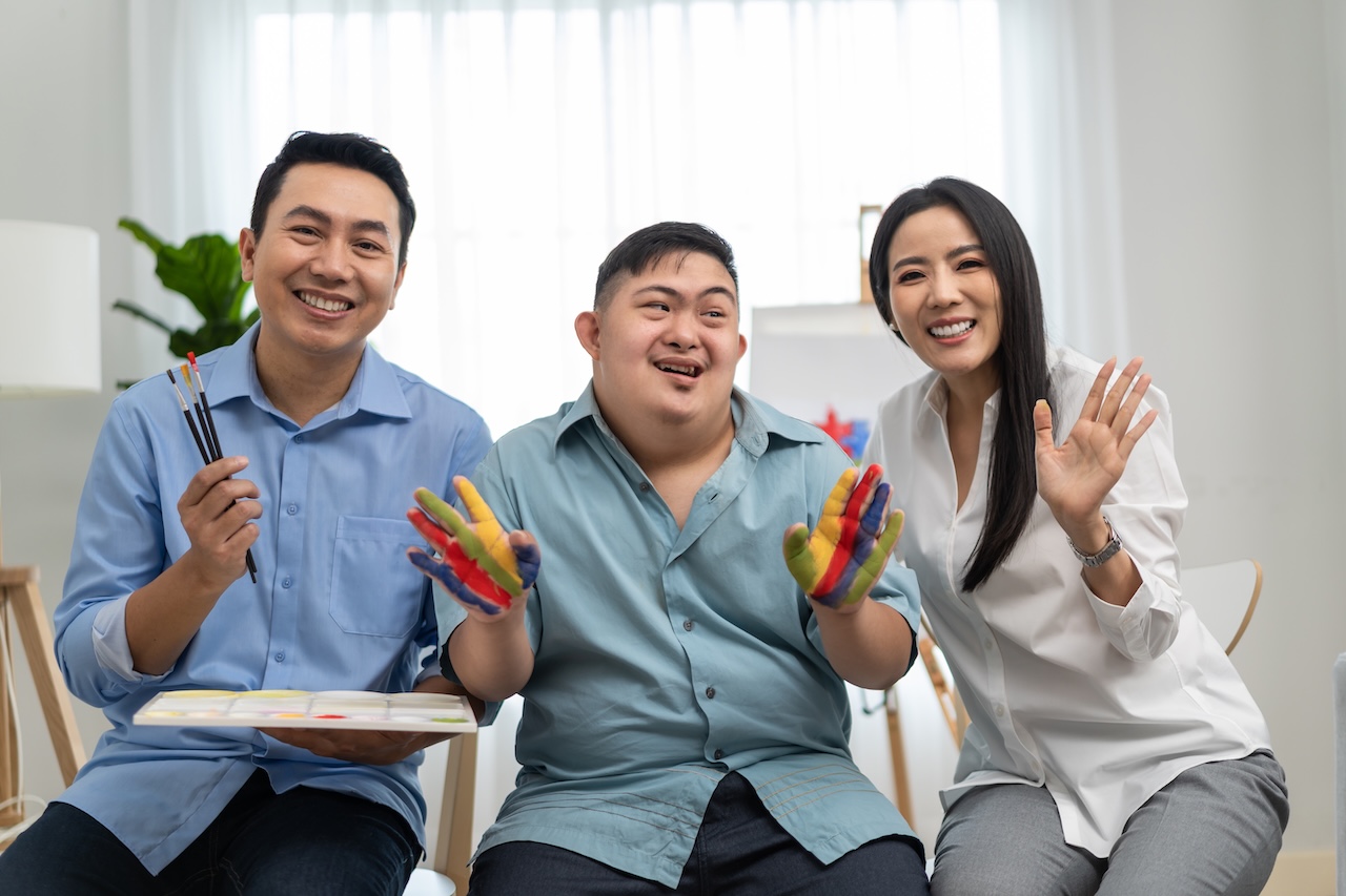 a young man with down syndrome is sat between his parents. the father, on the left, has paint and paintbrushes in his hands. the mother, on the right, is waving. the young man in the middle has his hands painted in a striped pattern of blue, red, green and yellow. they are all looking forward and smiling.