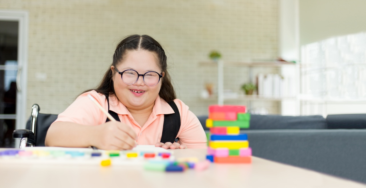 a young woman with down-syndrome is sat at a table indoors. she has glasses on and is in a wheelchair. she is painting. in front of her, there are colourful blocks stacked up. she is smiling.