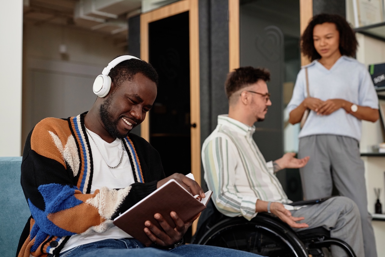 a group of people are standing around in an office setting. two people on the right are in discussion with each other, one is a wheelchair-user. the man in focus is sat on a sofa wearing headphones, and smiling down at a notebook as he writes in it.