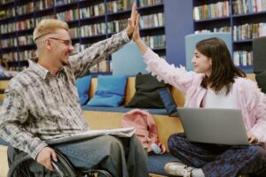 a woman and a man are sat in a library. the man, on the right, is in a wheelchair, with a notebook in his lap. the woman, on the left, is sat cross-legged on a sofa, with a laptop in her lap. they are both looking at each other and smiling. they are high-fiving.