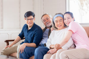 a family are sitting on a sofa together indoors. the parents are sat in the middle, the son is on the left, the daughter is on the right. they are all hugging and smiling. the parents are elderly.