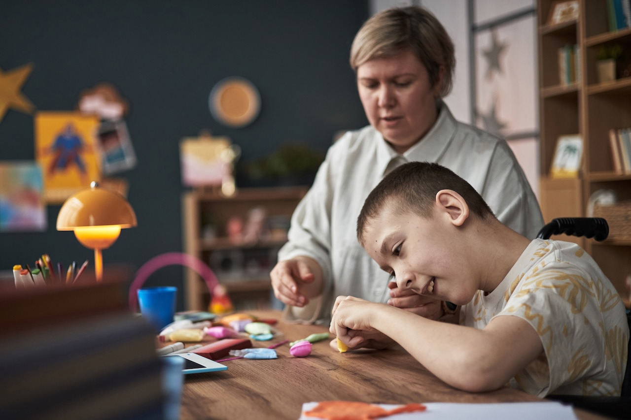 a young boy and his mother are sat at a table indoors. the boy in a wheelchair appears to be playing with some kind of putty. he is holding yellow putty and smiling. his mother is sat next to him with her hands out. she looks concentrated.