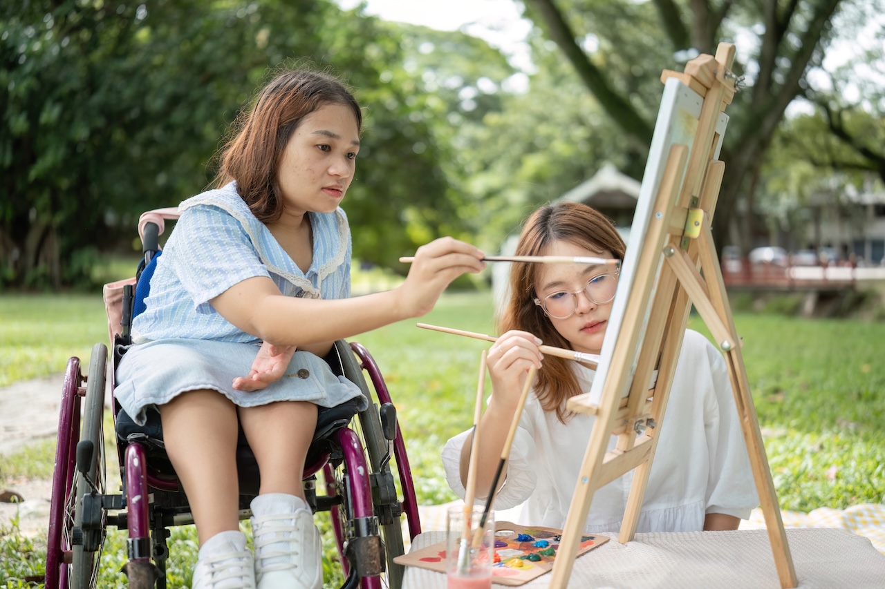 two young women are sat outside on the grass. the woman on the right is sat down on a blanket. the woman on the left is in a wheelchair. in front of them there is an easel with a canvas, a board with paint, and a glass with paint water. they are both painting on the canvas with brushes. they both look concentrated.