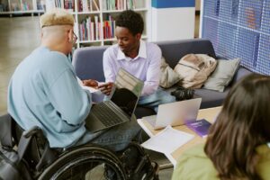 two men are sat around a table in a library. they are both looking down at something on a piece of paper. the man on the right is sat on a sofa. the man on the left is in a wheelchair. he has a laptop in his lap.