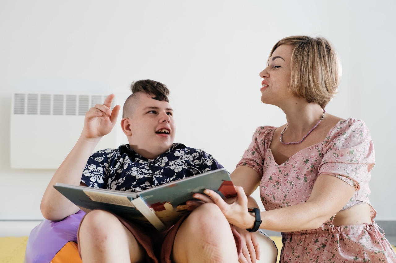 a young boy and his mother are sat indoors. he is sat in a colourful beanbag chair, with a book in his lap. he is holding one hand up and looking up at his mother. the mother is holding the book with and looking down at him. they are both smiling.