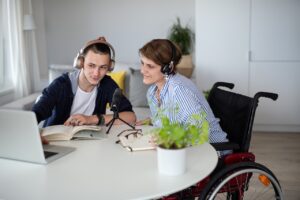 two people are sat at a table indoors. they are both wearing headphones. the person on the right is a wheelchair-user. they are both looking closely at a laptop, with notebooks out in front of them. there is also a microphone in front of them, as if they are recording something.