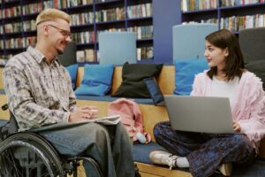 a man and a woman are sat across from each other in a library. the man is a wheelchair user, and has a notebook in his lap. the woman is sitting cross-legged with a laptop in her lap. they are both smiling.
