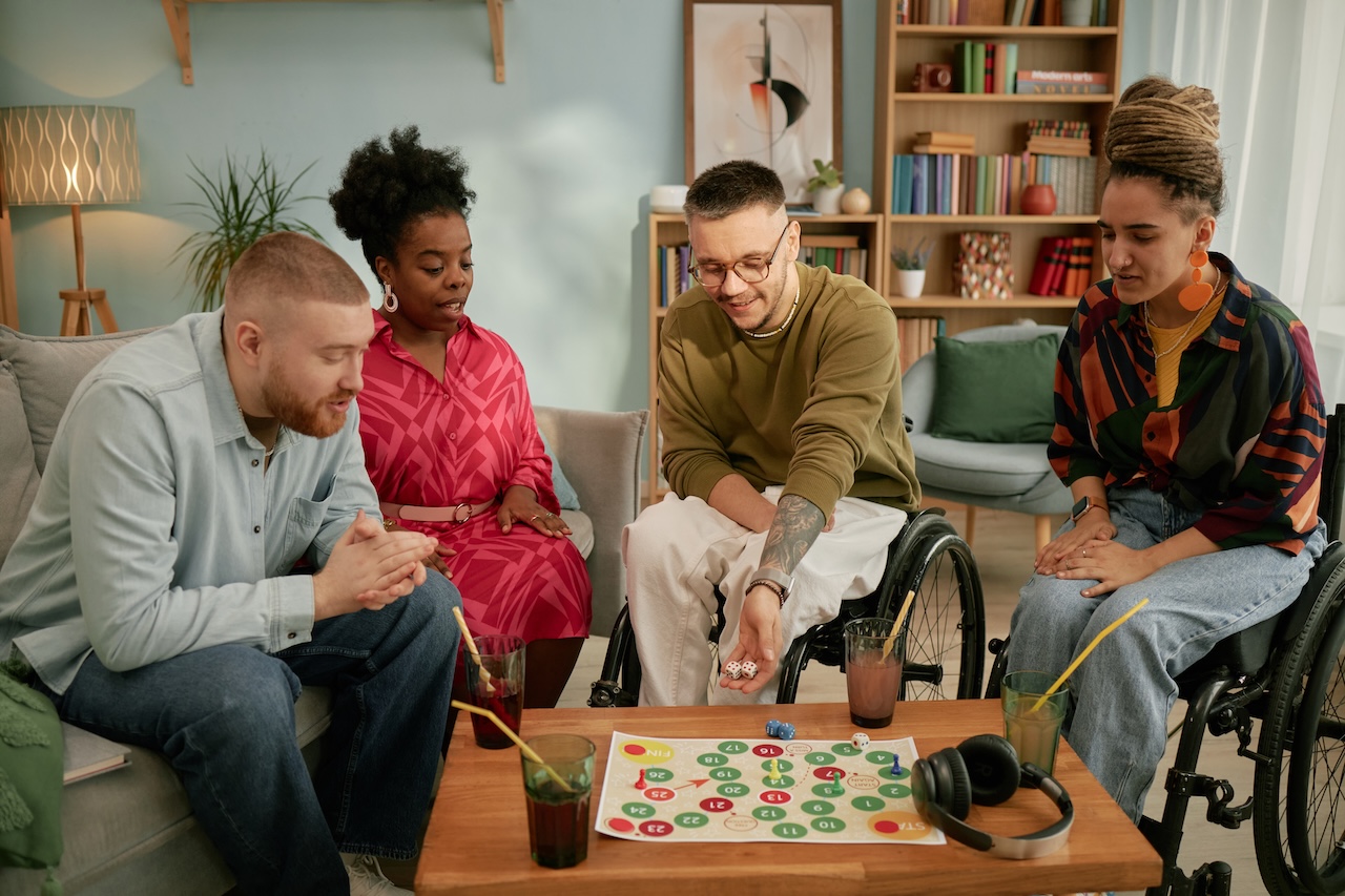 a group of young adults are sat indoors around a coffee table. on the coffee table there is a board game, headphones, and four drink glasses with straws. the two people on the left are sat on a sofa. the people on the right are both in wheelchairs. the man on the right is rolling the dice on the table. they all look concentrated on the game.
