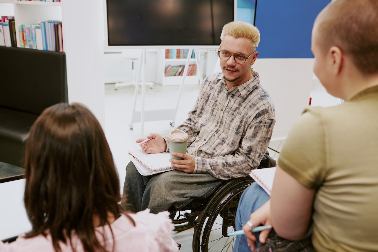 a group of three people are sat together in an office-setting. only the back of two of them are visible. the man in focus is in a wheelchair. he is holding a coffee cup and a pen. in his lap there is an open notebook. he seems to be discussing something with the other two people. behind him there is a large tv monitor.