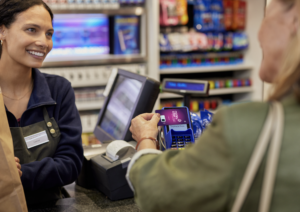A woman's hand is tapping a purple Biometric Fingerprint Card from Money Carer against a payment terminal in a shop. The cashier is looking at her and smiling.