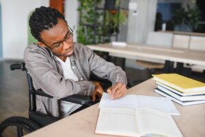a young man in a wheelchair is sat at a desk indoors. on the desk in front of him there is a stack of books, and two open notebooks. he is writing in the notebook closest to him. he looks concentrated.