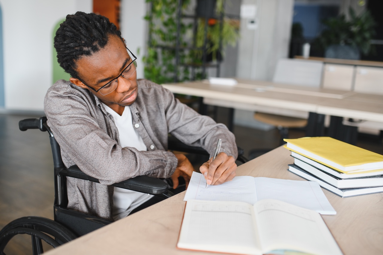 a young man in a wheelchair is sat at a desk indoors. on the desk in front of him there is a stack of books, and two open notebooks. he is writing in the notebook closest to him. he looks concentrated.