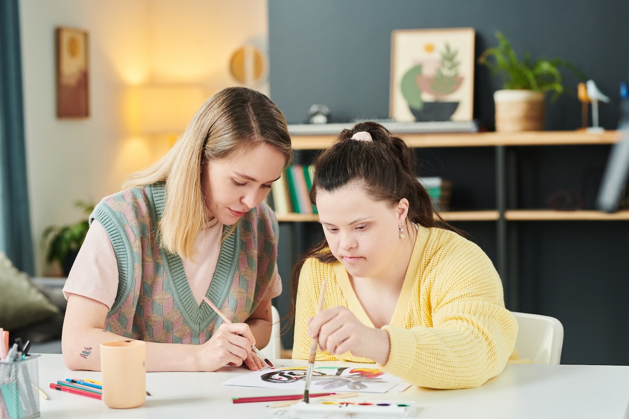 two young women are sat at a table indoors. they are painting on a piece of paper in front of them. the woman on the left is painting on the paper. the woman on the right is dipping her paintbrush into the paint. they both look concentrated.