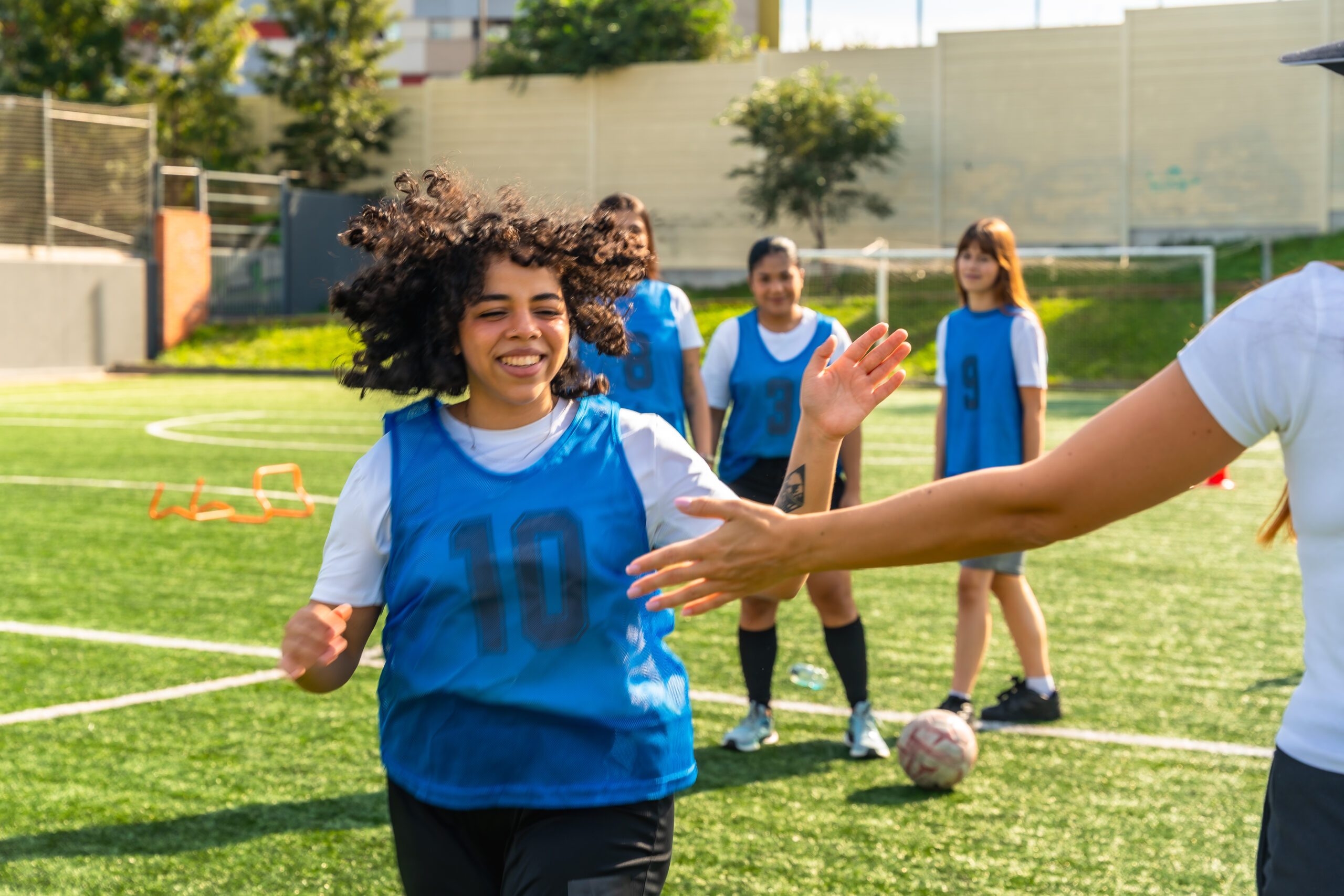 a group of young women are outside on a football pitch, on a sunny day. they are wearing blue sports jerseys. the young woman in focus is running forwards towards an outstretched hand, as if to give a high-five. they are all smiling.