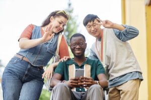three young people are outside. the man in the middle is in a wheelchair. he is holding his phone in front of him as if taking a picture. the man to his right is holding a peace sign and looking down at the camera. the woman to his left is waving and looking down at the camera. she has sunglasses on her head. they are all smiling.