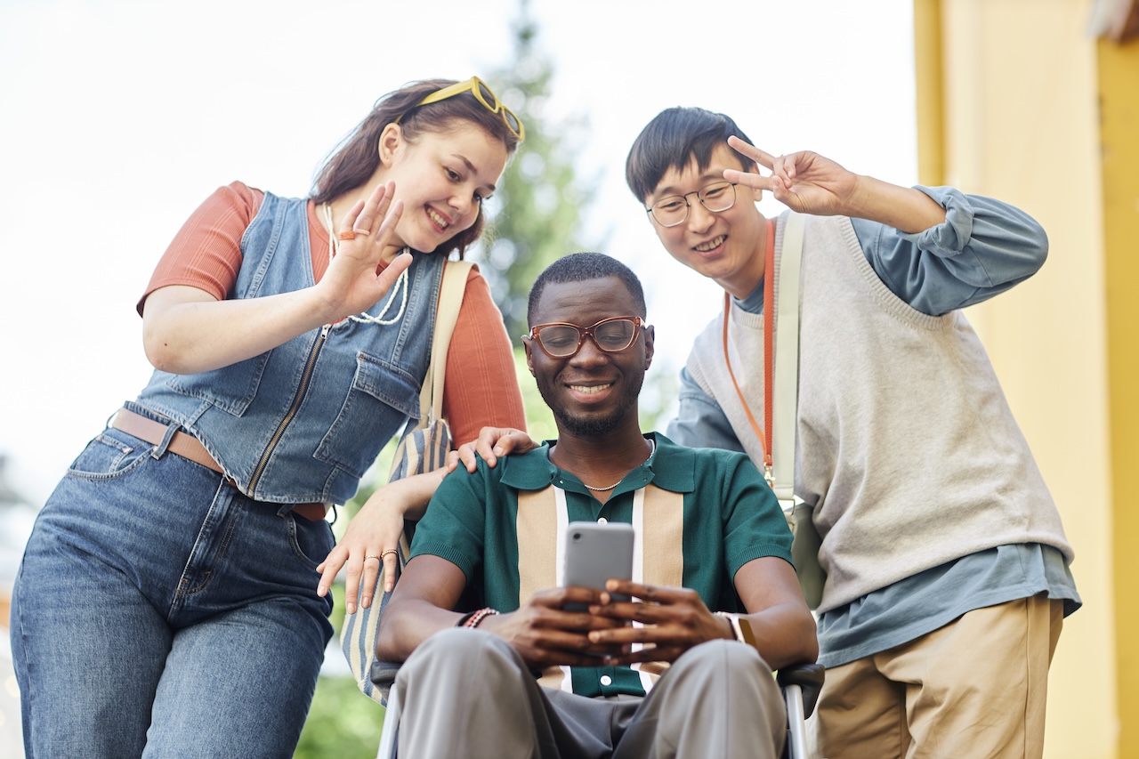 three young people are outside. the man in the middle is in a wheelchair. he is holding his phone in front of him as if taking a picture. the man to his right is holding a peace sign and looking down at the camera. the woman to his left is waving and looking down at the camera. she has sunglasses on her head. they are all smiling.
