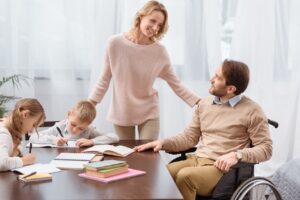 a family are sat at a table. the father, on the right, is in a wheelchair. he is looking up at the mother who is stood up. they are both smiling. she has her arm on his shoulder. the son and daughter, on the left, are doing their homework. they both look focused. the mother has her arm on the back of the son's chair.