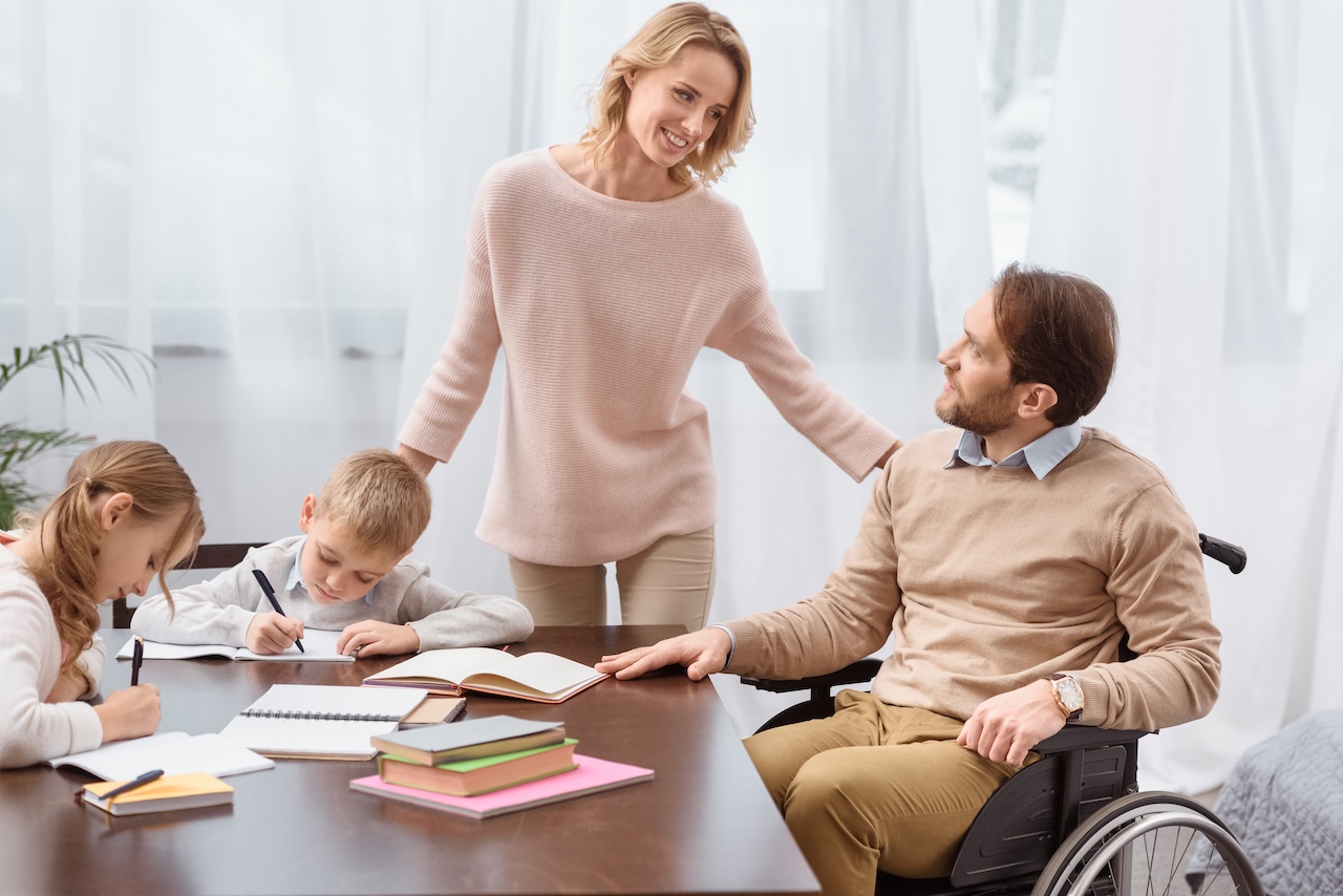 a family are sat at a table. the father, on the right, is in a wheelchair. he is looking up at the mother who is stood up. they are both smiling. she has her arm on his shoulder. the son and daughter, on the left, are doing their homework. they both look focused. the mother has her arm on the back of the son's chair.