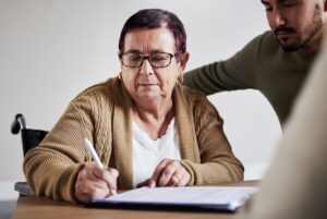 a woman in a wheelchair is sat at a table signing a piece of paper. there is a young man stood next to her with his arm around her shoulders. they are both looking down at the piece of paper. they both look concentrated.